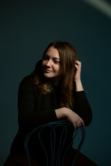 Classic dark studio portrait of a young brown-haired woman in black clothes sitting on a chair.