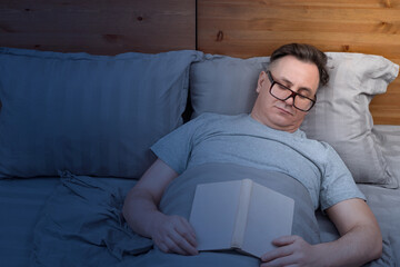 A man with glasses sleeps at night in bed with a book in his hands by the light of a night light
