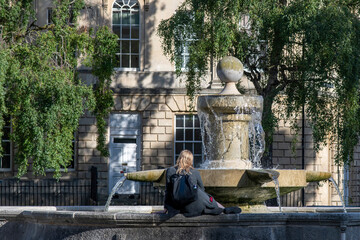 Close up view of a blond headed girl with bag on her back sitting on the edge of the water basin...