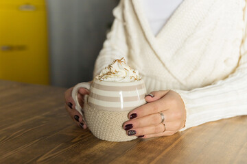 Morning coffee with whipped cream. winter composition. Hand with a cup of Cappuccino or latte coffee on christmas lights background.mug of hot cocoa in the hands of a girl with a dark red manicure