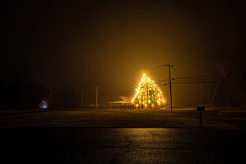 Broomes Island, Maryland USA A large glowing and illuminated tree with Christmas lights in the fog on a rural road in the mist at night. © Alexander