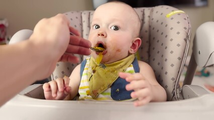 baby eats dirty. mom feeds with a spoon in the table for feeding baby boy. dirty baby messy kid lifestyle eats from a spoon indoors. kindergarten kids dream concept