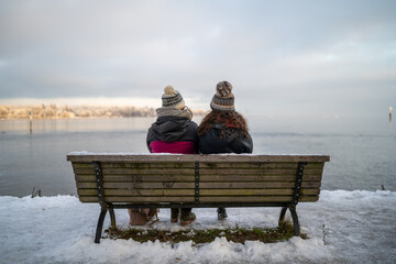 Two friends sitting next to each other on a bench in winter looking out onto a panoramic lake