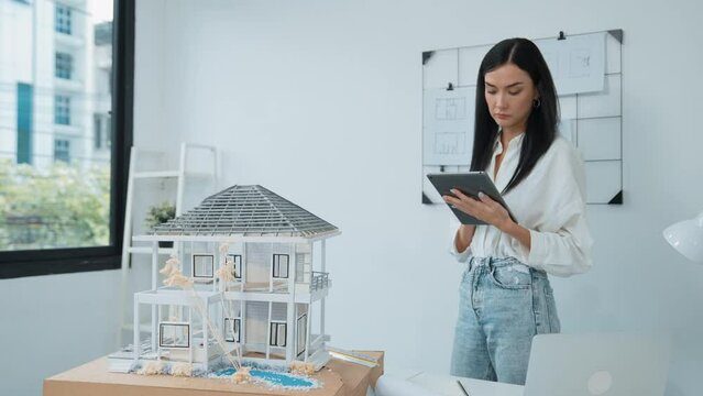 Beautiful caucasian architect focus on using tablet to check house model on meeting table with blueprint, house model and architectural document scatter around at meeting room. Immaculate.