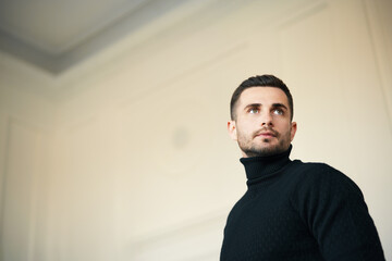 Pensive man in a black turtleneck looking upward in a spacious, light-filled room.