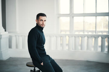 Trendy handsome man sits thoughtfully on chair in spacious white room