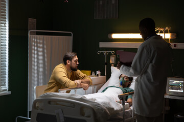 Beside the unconscious girl wearing an oxygen mask, a man is seated, while an African doctor...