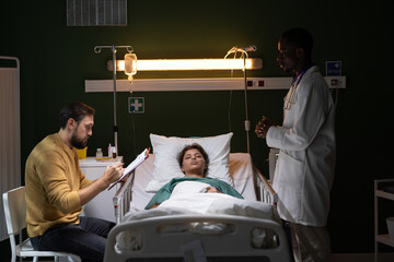 In hospital room, man sits beside bed, attentively examining consent form, signs unconscious wife...