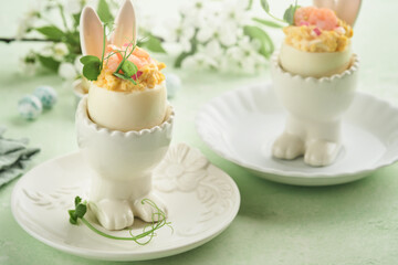 Stuffed or deviled eggs with yolk, shrimp, pea microgreens with paprika in rabbit-shaped stand for easter table decorate fresh cherry or apple blossoms on light background. Traditional dish for Easter