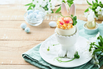 Stuffed or deviled eggs with yolk, shrimp, pea microgreens with paprika in rabbit-shaped stand for easter table decorate fresh cherry or apple blossoms on light background. Traditional dish for Easter