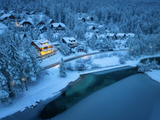 Aerial view of fairy village in snow, road, forest, Jasna lake, houses, street lights at winter night. Top view of alpine mountains, illumination, snowy pine trees at twilight. Kranjska Gora, Slovenia