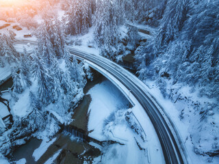 Aerial view of bridge, river with waterfalls in snowy fairy forest in winter at sunset. Top drone view of mountain road, pine trees in snow at dusk. Beautiful rural road in woods. Travel in Slovenia
