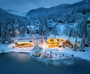 Aerial view of illuminated houses in fairy village in snow, forest, Jasna lake, street lights at winter night. Top view of alpine mountains in fog, snowy pine trees at dusk. Kranjska Gora, Slovenia - obrazy, fototapety, plakaty