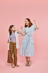 full length of carefree and trendy woman with preteen daughter dancing on pink backdrop in studio