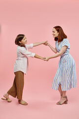 full length of carefree and trendy woman with preteen daughter holding hands and dancing on pink