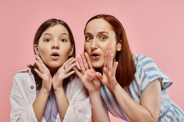 surprised mother and daughter holding hands near open mouths and looking at camera on pink