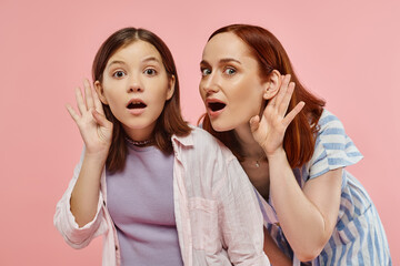 curious mother and teenage daughter holding hands near ears and eavesdropping on pink backdrop