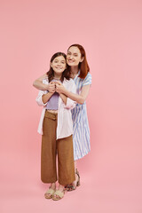 joyful woman embracing teenage daughter and holding her hands on pink backdrop, full length
