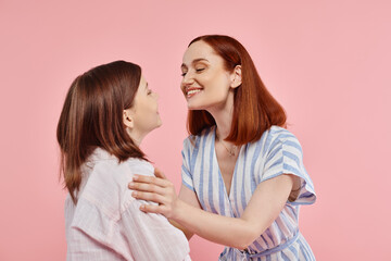 joyful and stylish woman with teenage daughter smiling at each other on pink backdrop in studio