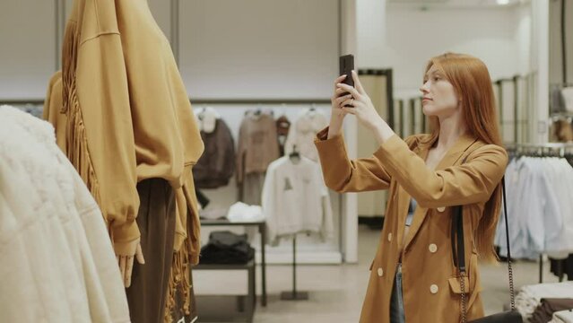 Medium shot of young beautiful ginger haired woman texting and taking pictures on smartphone of stylish camel sweatshirt with fringes on mannequin in fancy designer showroom