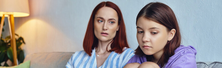 worried and caring mother looking at depressed teenage daughter on couch in living room, banner