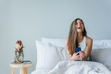 Excited young brunette woman sitting in bed holds phone laughing loudly eyes closed happy, received...