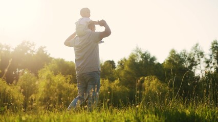 father carries a little son on his shoulders and walks on the grass in the park. happy family childhood dream concept. a small child sits on the shoulders of his dad man walking in lifestyle field