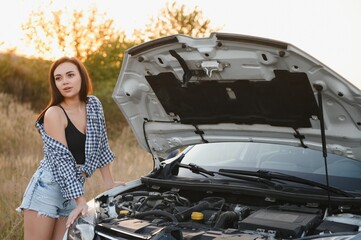 Beautiful slim girl in shirt and shorts looks in open car hood on a road