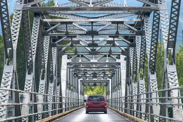 Drivers perspective on a steel truss bridge across the river in Canada with going red van driving in front on bridge deck of steel grating - obrazy, fototapety, plakaty