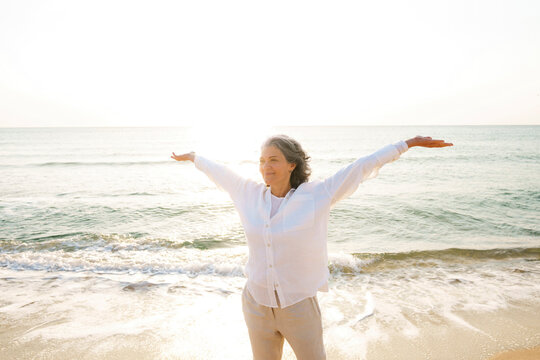 Smiling mature woman with arms outstretched standing at beach on sunny day