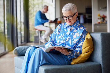 Senior woman sitting on couch at home reading magazine