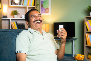 middle aged man laughing while watching comedy show on tv or television by holding remote at home -...