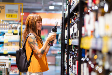 Side view of smiling young tanned Caucasian woman with tattoo choosing bottle of vine. Shopping and consumerism. Copy space