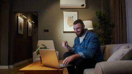 Bearded young adult man sitting on sofa while using laptop checking messages read notice receive...