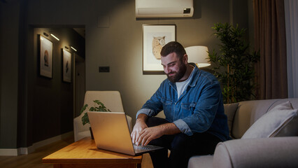 Young bearded man sitting and relaxing on sofa at home using laptop enjoying surfing internet,...