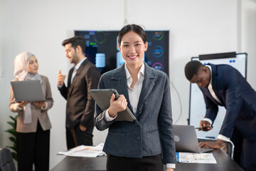 portrait of young asian businesswoman in formal suit with tablet standing in the international office,  group of multiracial team colleague diverse employee coworker meeting for financial performance