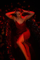 Fototapeta na wymiar red light, sensual young woman in slip dress lying in black bathtub among beautiful flowers