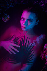 fashion shot, charming woman lying among palm leaves and flowers in water, blue and purple lights