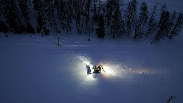 Aerial view of working snowcat on the snowy alpine ski slope at winter night in Kranjska Gora, Slovenia. Top drone view of snowcat grooming snow, ski resort, lights at dusk. Snowplough machine
