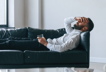 Having headache, lying down on the sofa. Upset young man drinker alcoholic is indoors