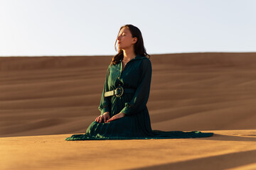 woman in green long dress stands in desert sand dunes background yellow orange sunset