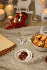 Bowl of cookies, cup of tea, dry oranges, pine cones, books, reading glasses, small presents, various Christmas decorations and lit candles on the table. Cozy Christmas hygge. Selective focus.