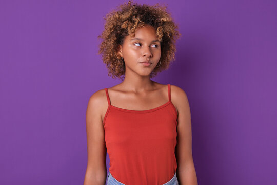 Young pretty curly African American woman teenager college student looking away from camera as sign of mistrust or watching people passing by stands on isolated purple background. Casual female pupil
