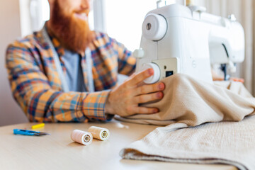 handsome redhaired man with long beard sews at a sewing machine at home studio