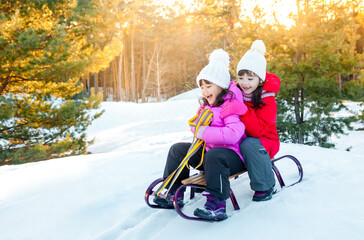 Happy funny children ride sleds on snowy road in forest. Family on winter walk. Two little girls are sledding down a hill. Children ride in the snow. Place for text.