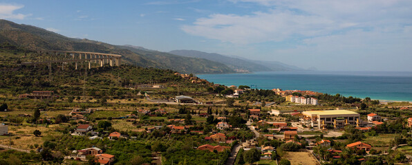 Picturesque panoramic landscape view of Sicily at sunny day. Curved automobile viaduct in mountains near Santo Stefano di Camastra. Small houses near the sea. Travel and tourism concept. Sicily, Italy