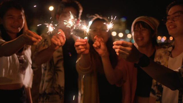 outdoor new year party and camping activity concept.Group of young Asian friends holding sparkling fireworks,lit light sparkler,dance together at beach.Party people, love friendship relationship.