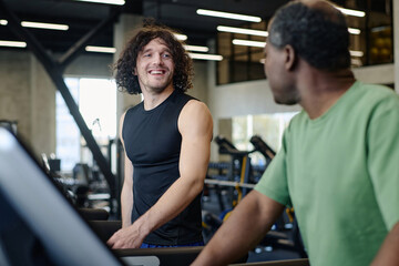 Fototapeta na wymiar Caucasian guy and african american man looking at each other walking on treadmill at gym