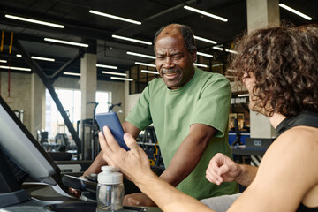 Elderly black man standing on treadmill at gym with young caucasian guy showing him his smartphone