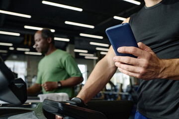 Unknown caucasian guy training at gym holding smartphone, elderly black man exercising on background